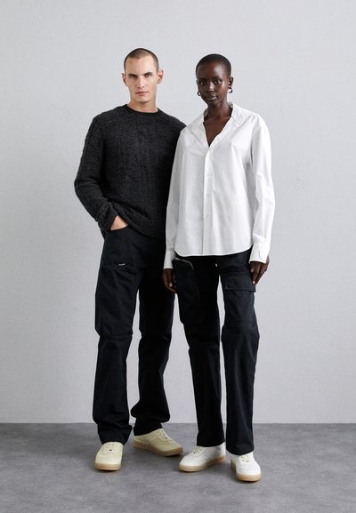 Two models stand side by side. The man wears a textured dark sweater and black cargo pants, while the woman wears a white button-up shirt and black pants.