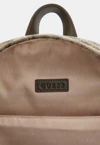 Interno di uno zaino in tessuto beige con fodera marchiata e una patchetta in pelle marrone. Presenta una texture liscia e un design semplice e pulito.