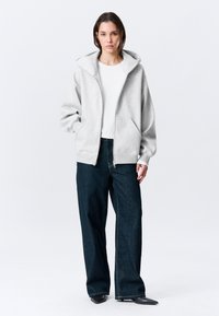 Grå hettejakke med glidelås, romslig hette, lange ermer og frontlommer, kombinert med løstsittende mørke denimjeans. Enkel design, minimal tekstur.