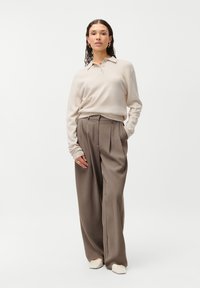 Camisa polo bege de manga longa com colarinho; combinada com calças largas plissadas de tom castanho claro; estilizada com sapatos planos de cor clara.