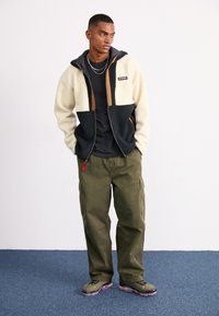 Fleece zip-up jakke i creme og marineblå med brune detaljer; sort T-shirt under; olivengrønne cargo-bukser; grønne og sorte sneakers.