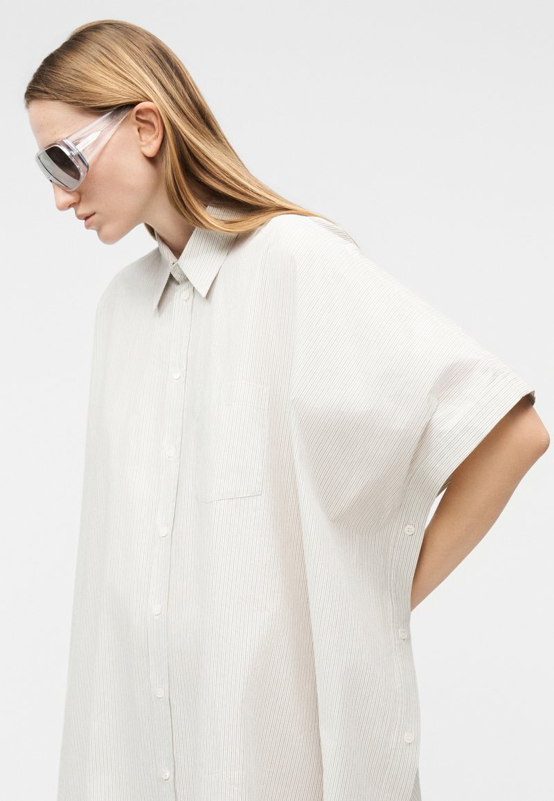 Femme aux longs cheveux blonds portant une chemise à rayures beige clair surdimensionnée et des lunettes de soleil à monture transparente, regardant vers le bas, sur un fond blanc uni.