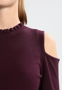 Bordeauxfarbener Pullover mit elastischem, gerüschtem Ausschnitt und Cutouts an den Schultern. Weicher Stoff mit tailliertem Design und kurzen Ärmeln.
