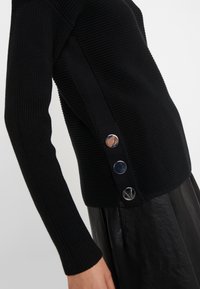 Pull noir à manches longues côtelé avec trois grands boutons argentés sur le côté, porté avec une jupe en cuir noir.