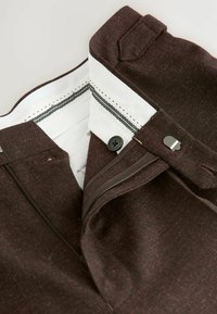 Pantalon en tissu texturé marron avec une doublure blanche, comportant une fermeture éclair et un bouton. Inclus des coutures contrastantes et une poche latérale.