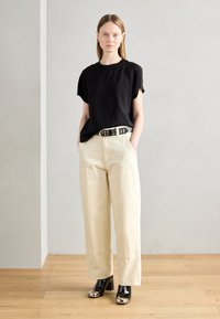 T-shirt noir à manches courtes, pantalon large crème, chaussures noires avec des détails métalliques, ceinture argentée cloutée, silhouette droite, design minimaliste.