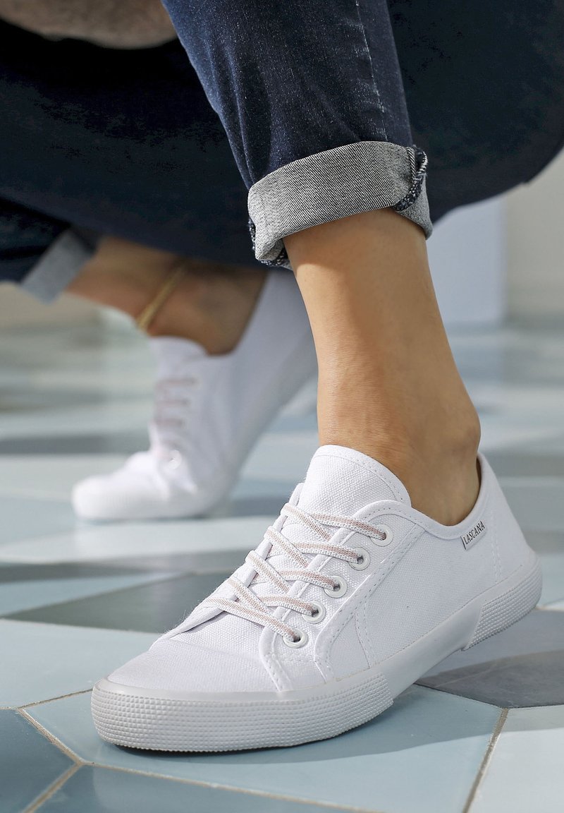 White canvas sneakers with round toe, lace-up design, and rubber soles. The shoes are paired with rolled denim pants showcasing a clean look.