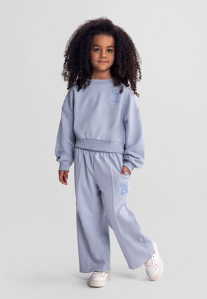 Lapsi, jolla on kiharat hiukset, pukeutuneena vaaleansiniseen, kirjailtuine kuvioineen olevaan collegepaitaan ja housuihin, seisoo yhdellä kädellä taskussa.