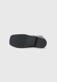 Suola nera di scarpa con design piatto, caratterizzata da una superficie in gomma testurizzata, scanalature verticali e forma arrotondata. Estetica minimalista e materiale resistente.