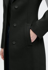 Hosszú fekete kabát sima anyagból, nagy fekete gombokkal, oldalzsebbel és szabott ujjakkal.