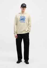 Felpa beige con stampa grafica blu e logo, abbinata a pantaloni cargo neri con tasche laterali e scarpe nere. Cappello nero con logo.