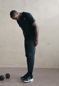 Czarny T-shirt Nike i ciemnozielone dresy noszone przez mężczyznę. Czarne buty sportowe z białymi akcentami. Hantla na ziemi obok.