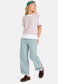 Un gilet sans manches en tricot côtelé rose clair par-dessus un t-shirt blanc, associé à un pantalon en tartan vert et des chaussures à enfiler marron.