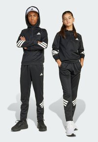 Ropa deportiva negra que incluye una sudadera con cremallera y pantalones de chándal. Ambos tienen rayas blancas en las mangas y las piernas, y están hechos de una tela suave.