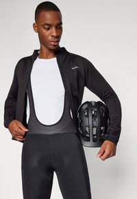 Svart sykkeljakke med lange ermer, hvit basislag t-skjorte, svarte bib-shorts og en svart hjelm med ventilasjonsåpninger.
