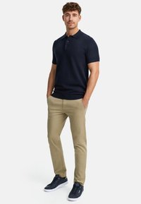 Polo-shirt à manches courtes bleu marine, pantalon beige et baskets noires. Le polo présente un col et une patte de boutonnage à trois boutons, mettant en valeur une coupe ajustée.