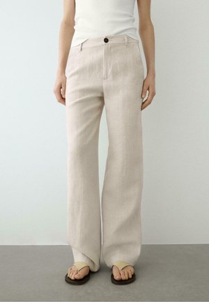 Vrouw draagt beige wijde broek met een zwarte knop, wit mouwloos topje en beige sandalen, staand op een grijze vloer.