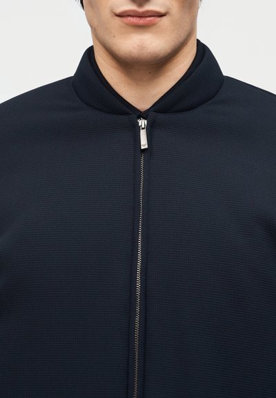 Κοντινό πλάνο ενός ατόμου που φοράει ένα υφασμάτινο σκούρο μπλε navy σακάκι με ασημένιο φερμουάρ και ribbed γιακά.