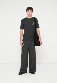 Μαύρο βαμβακερό t-shirt με λογότυπο, συνδυασμένο με γκρι παντελόνι φαρδύ στο πόδι. Το μοντέλο κρατάει μία μαύρη τσάντα με υφή και φοράει μαύρα παπούτσια. Μινιμαλιστικός σχεδιασμός.