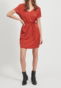 Robe à manches courtes et encolure en V, de couleur orange rouille, avec une ceinture à nouer à la taille, en tissu lisse, associée à des bottines noires.