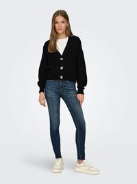 Cardigã preto em malha com três grandes botões metálicos, combinado com jeans skinny azul escuro e ténis chunky bege.