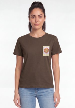 Femme aux cheveux foncés portant un t-shirt vert olive avec un patch graphique pizza devant et un jean bleu clair sur fond blanc.