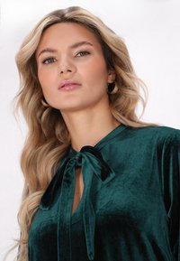 Top din catifea de culoare verde închis cu detaliu la gât sub formă de fundă. Materialul are o textură netedă și o ușoară strălucire. Modelul prezintă o vedere frontală cu accesorii minime.