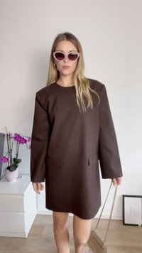 Smeđa oversized haljina s okruglim dekolteom, dugim rukavima, prednjim džepovima i glatkom teksturom. Uparena s ružičastim mačkastim naočalama i malom torbicom.