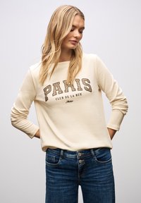 Kremowa bluza z napisem "PARIS" i "CLUB DE LA RUE" w kolorze brązowym, okrągły dekolt, długie rękawy. Noszona z ciemnoniebieskimi dżinsami.