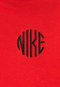 Tejido rojo de textura suave con el logotipo bordado en negro de "NIKE", estructurado en una orientación vertical dentro de una forma ovalada.