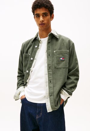Giacca camicia in corduroy verde con tasca frontale, T-shirt bianca sotto e jeans in denim scuro. Presenta chiusura con bottoni e dettagli a quadri.