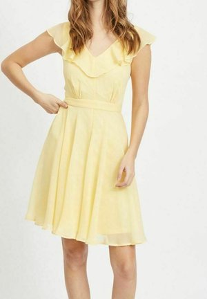 Mujer con un vestido amarillo sin mangas hasta la rodilla, con escote con volantes y cintura ceñida, de pie frente a un fondo blanco liso.