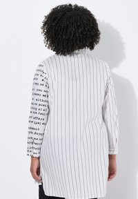 Blusa bianca con strisce nere verticali e un motivo testuale nero su una manica. Presenta maniche lunghe e un taglio diritto.