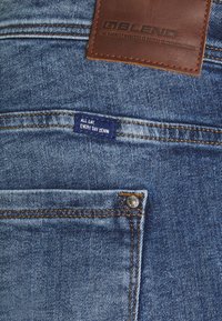 Blå denim jeans med ljus tvätt, med en läderbakpatch, orange sömmar och en etikett som står "All Day, Every Day Denim."