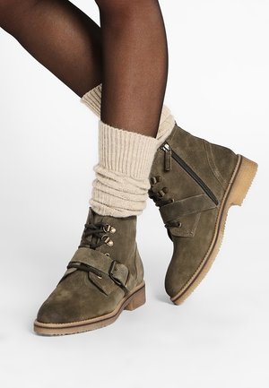 Bottines en daim vert olive avec boucle, fermeture éclair et détails en dentelle, portées avec des guêtres en maille beige et des collants noirs transparents.