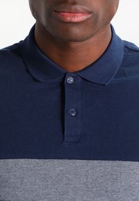 Homme portant un polo bleu marine avec deux boutons et une section à rayures horizontales gris clair sous la poitrine.
