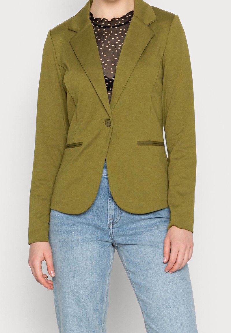 Olivově zelený blazer s jemnou texturou, jedním knoflíkem, klopovými límci a dvěma předními kapsami, spárovaný s světle modrými džínami.