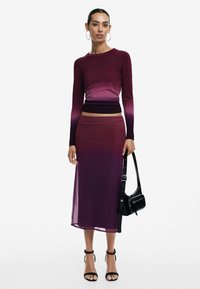 Langärmliges Gradient-Oberteil, das von dunkellila zu helllila übergeht, kombiniert mit einem passenden transparenten Midikleid und accessoriert mit einer schwarzen Tasche.