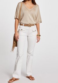 Beige metalen top met korte mouwen, witte flared jeans met voorzakken, een bruine riem en beige sandalen, aangevuld met een beige tas met franjes.