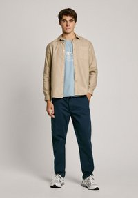 Camicia button-up tan sopra una t-shirt azzurra chiara, abbinata a pantaloni blu navy. Il modello indossa sneakers bianche; la camicia ha una tasca sul petto ed è leggermente slim fit.