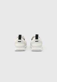 Λευκά αθλητικά παπούτσια με μαύρους βρόχους στη φτέρνα και μαύρο λογότυπο ζιγκ ζαγκ στη φτέρνα, από πίσω, σε απλό ανοιχτό γκρι φόντο.