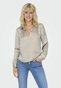 Blouse satin beige clair avec un col en V et des manches longues, associée à un jean bleu avec une fermeture à boutons et des poches avant.
