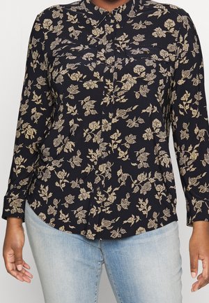 Sort skjorte med knapper med beige blomsterprint, lange ærmer og to brystlommer, båret med lyseblå jeans.
