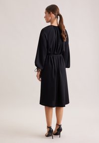 Schwarzes Kleid mit einem taillierten Gürtel, langen Ärmeln und einem runden Ausschnitt. Weicher Stoff, knielang und kombiniert mit schwarzen High Heels.