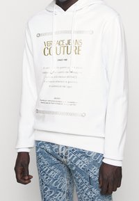 Vit hoodie med guldtext, med en framgrafik och snoddar. Parat med ljusblå jeans som visar upprepade logomönster.