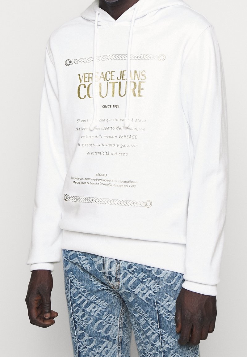 Vit hoodie med guldtext, med en framgrafik och snoddar. Parat med ljusblå jeans som visar upprepade logomönster.