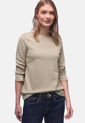 Jeune femme aux cheveux blonds portant un chemisier beige à manches longues et un jean bleu foncé devant un fond clair uni.