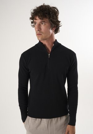 Zwarte geribbelde pullover met een rits bij de halslijn, met lange mouwen en een aansluitend ontwerp. Draagt over lichtgekleurde broek.