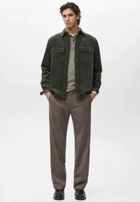 Hombre vestido con una chaqueta de camisa de pana verde oscuro, suéter beige, pantalones marrones y mocasines negros, de pie con las manos en los bolsillos.