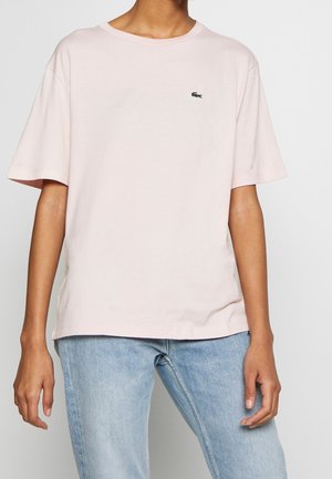 Jasnoróżowy bawełniany t-shirt z krótkimi rękawami, okrągłym dekoltem i małym zielonym logo Lacoste na lewej piersi. W zestawie z niebieskimi dżinsami.